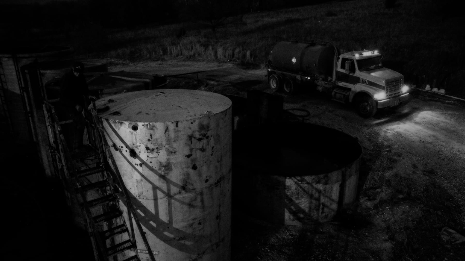 Oil storage tanks and a tanker truck at night, headlights burning.