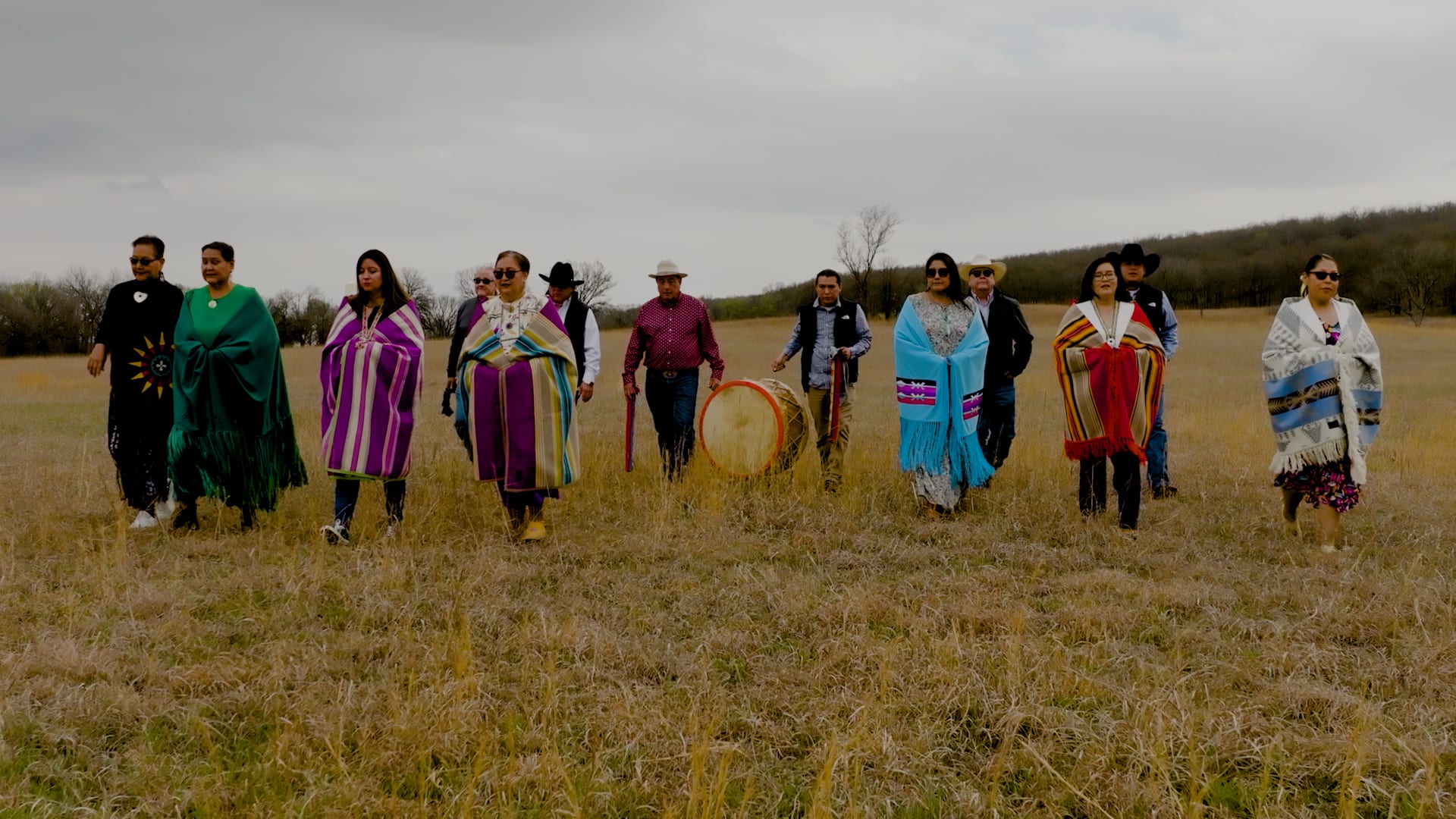 Osage citizens crossing the prairie in regalia, one carrying a large drum.