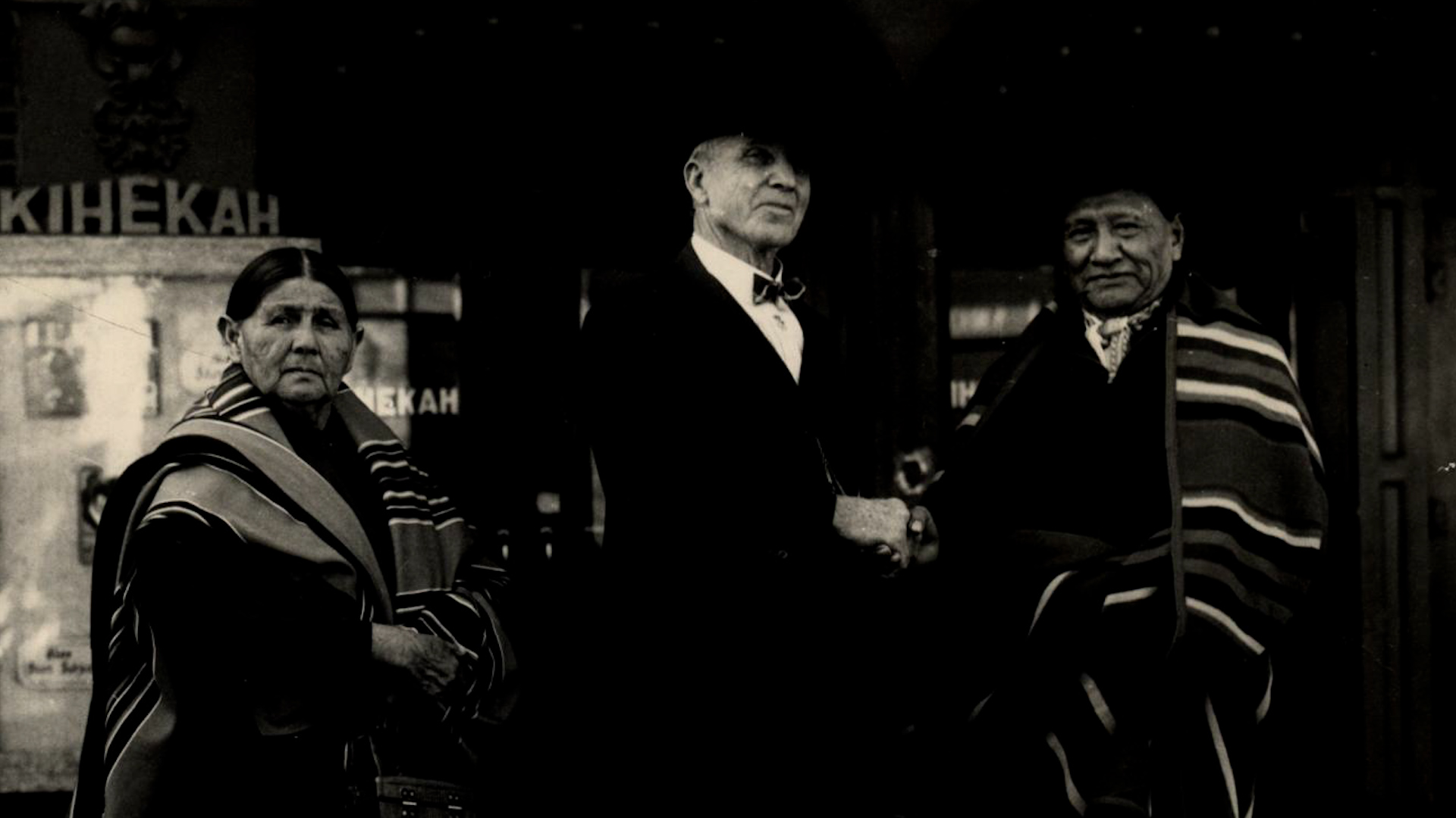 Three figures — two Osage elders and a white official — standing outside the Kihekah building.