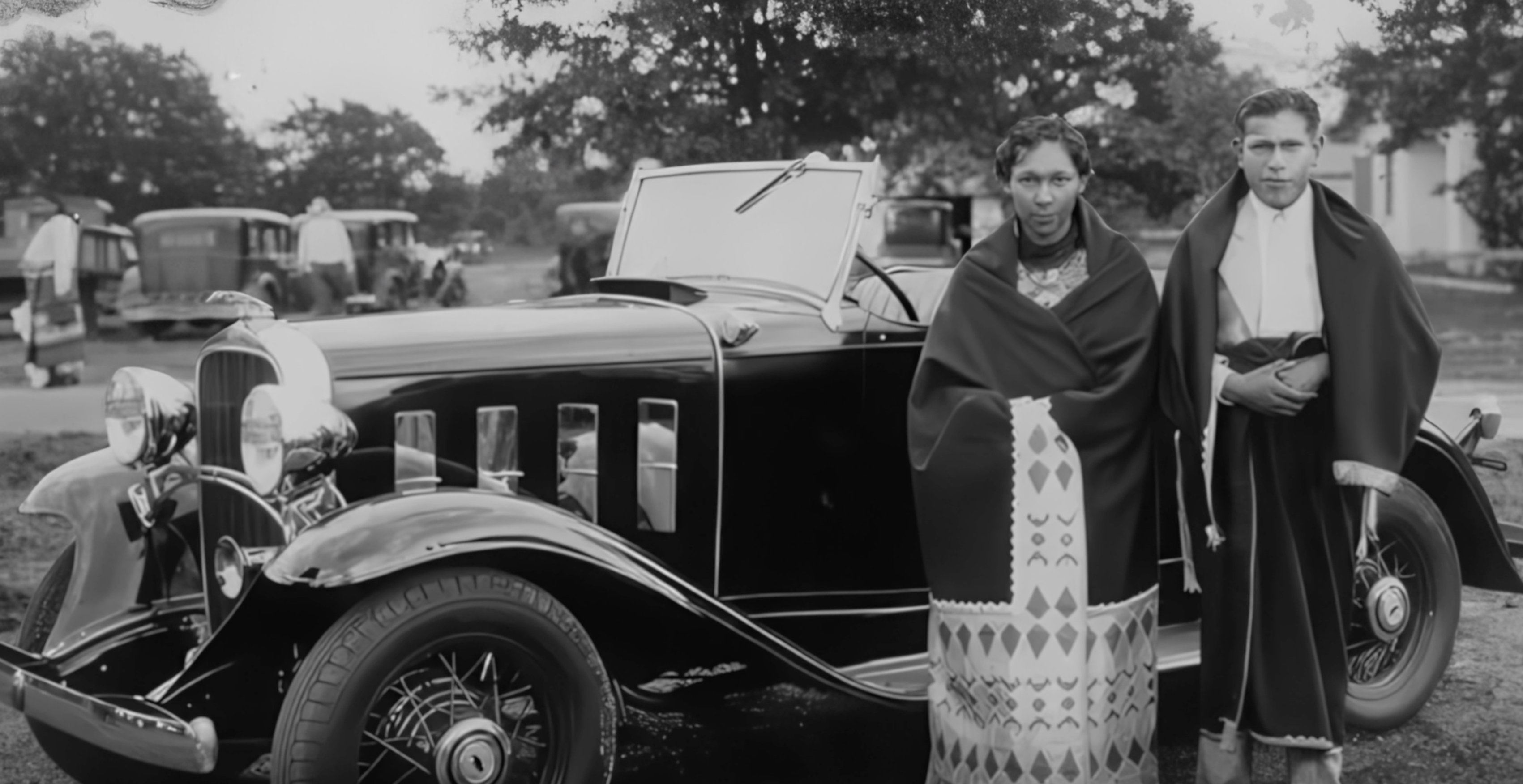 Two Osage members in traditional blankets beside a luxury automobile, circa 1920s.
